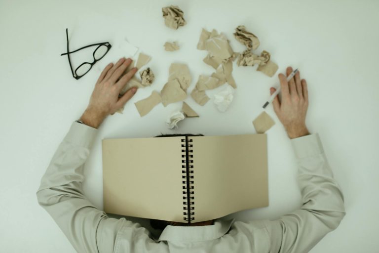 Tired man lying on the ground, glasses off, covering his head with a notebook while scattered sheets of paper surround him, symbolizing exhaustion and overwhelm.